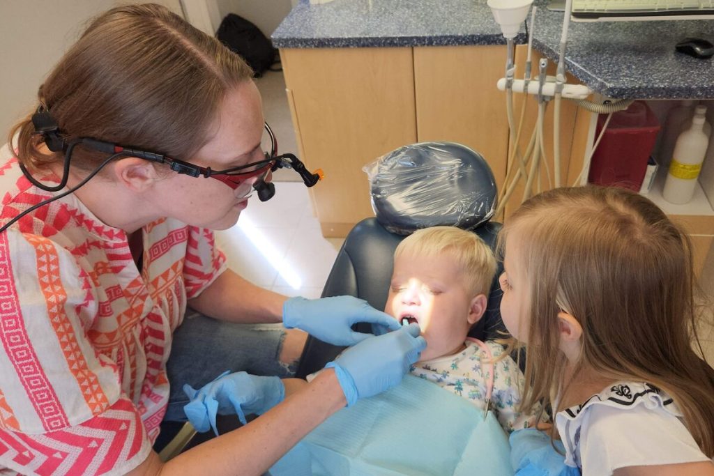 Dr. Barbosa, wearing magnifying loupes, checks a toddler's teeth while an older child leans in to watch