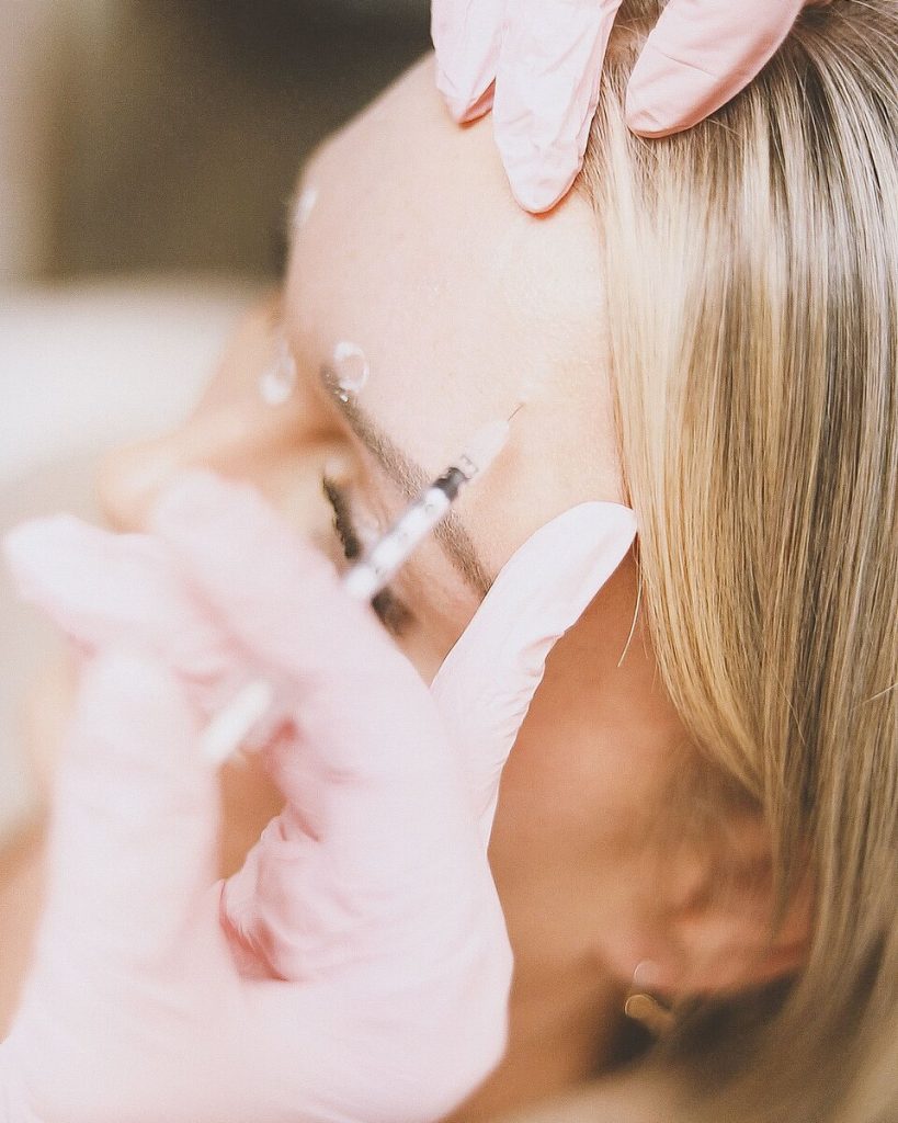 A gloved provider gives a small injection at the side of a reclining woman's forehead