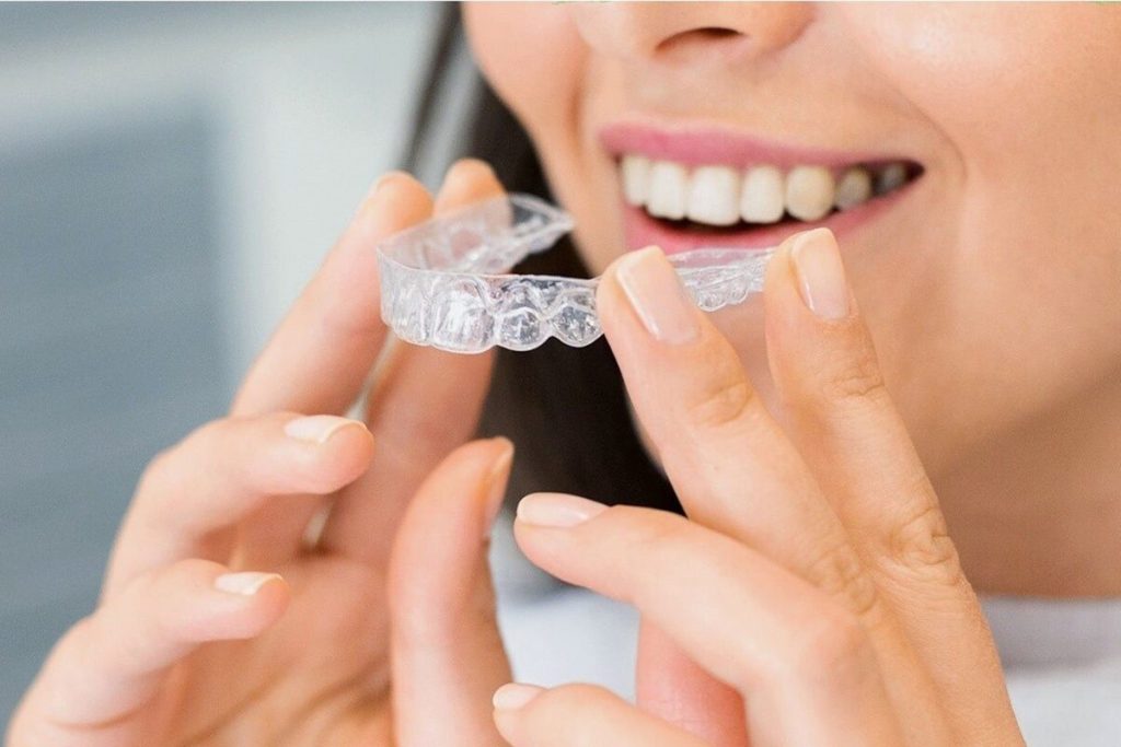 A smiling woman holds a clear aligner up beside her teeth